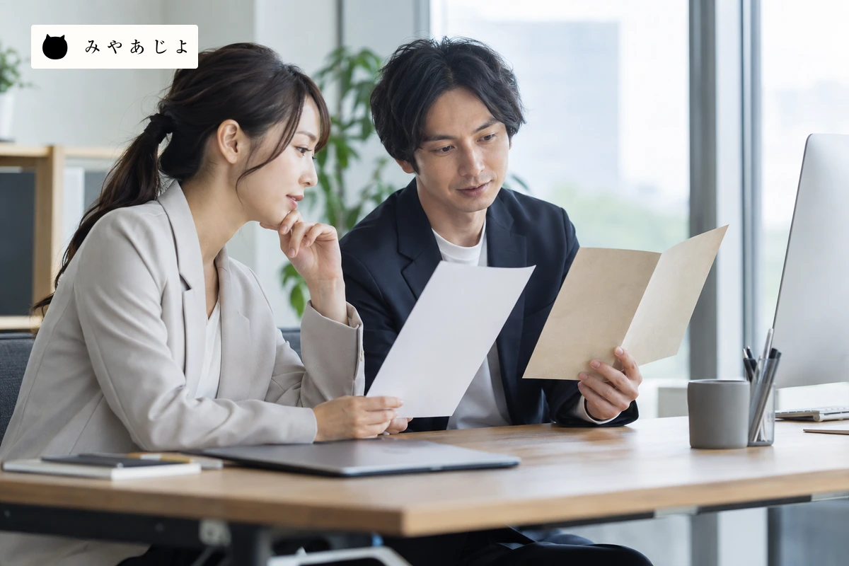 成人Web担当者二人が無地の旧版パンフレットと白紙の新資料を照らし合わせる場面