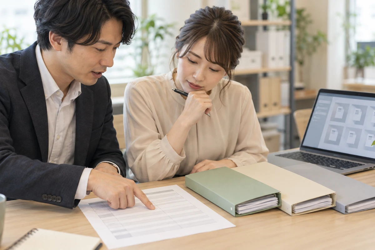オフィスの机で二人の担当者が無記名の資料一覧と空欄のチェック表を照合する場面