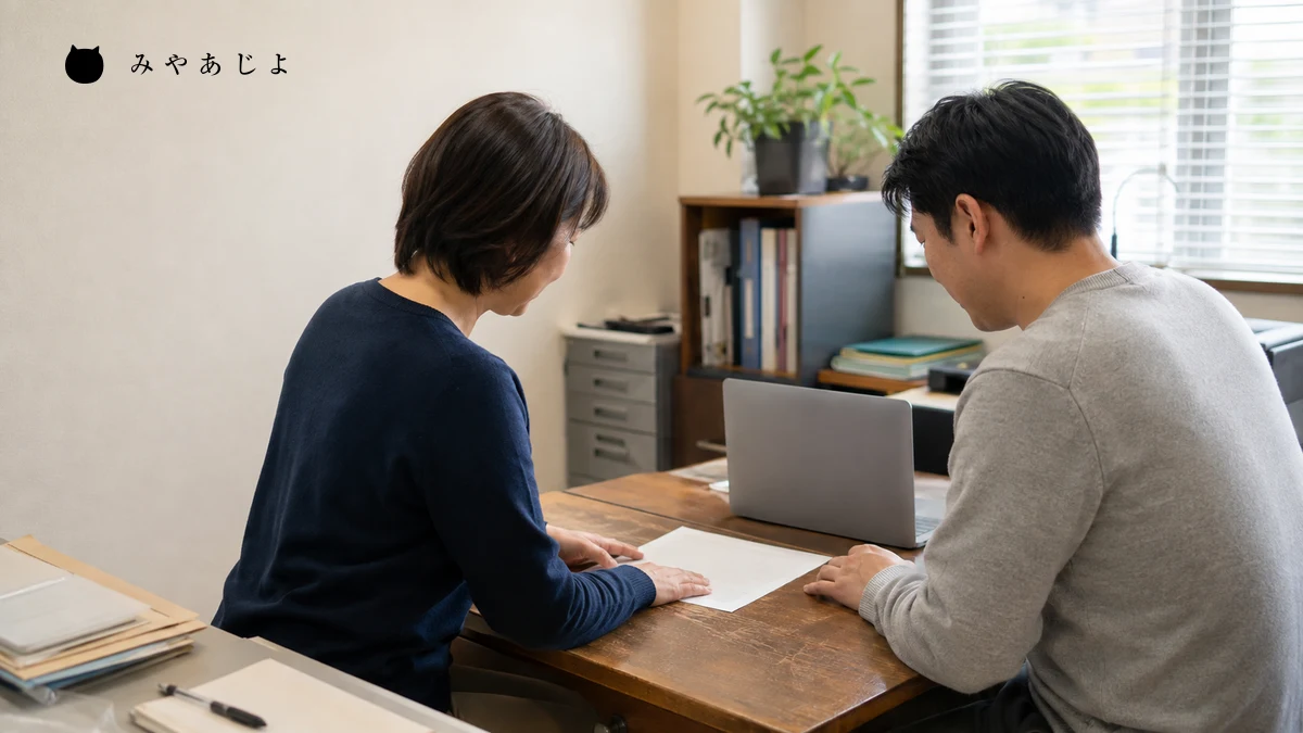 架空の小規模企業の二人の担当者が机上の案内資料と端末を使って代理更新の確認を行う編集イメージ